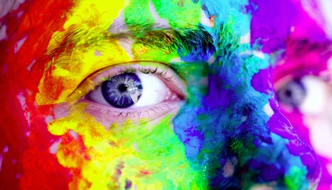 Close-up of a human eye surrounded by vibrant rainbow paint splashes and powder in red, yellow, green, blue, and purple hues.