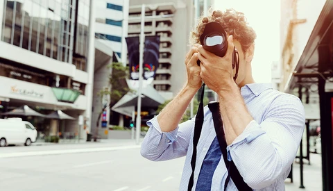 Photographer holding vintage camera up to face on urban street with tall buildings in background.