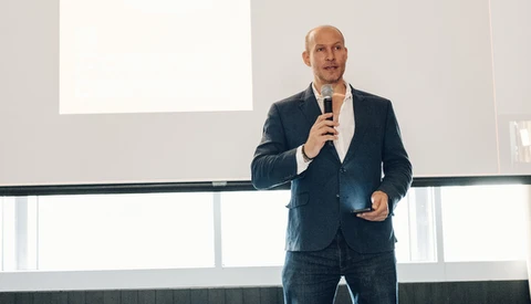 Bald man in dark blazer speaking into microphone during presentation with projection screen visible.