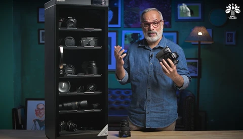 Man in denim shirt holding camera equipment while speaking in studio setting with gear display cabinet.
