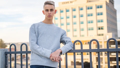 Young man in light blue sweatshirt leaning against railing with multi-story building behind him.