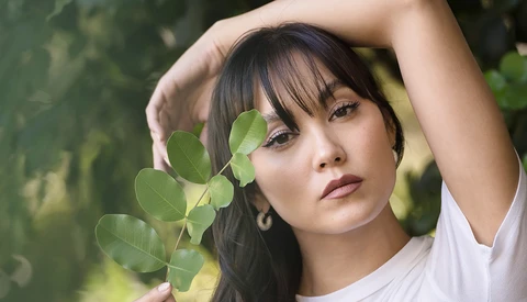 Portrait of a woman with dark hair holding green foliage near her face in natural sunlight.