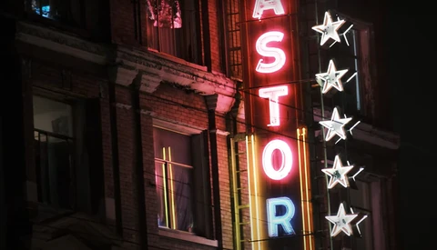Neon sign reading "ASTOR" with illuminated stars on a brick building facade at night.