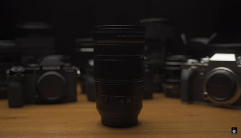 A telephoto lens in sharp focus sits centered on a wooden surface, with camera bodies blurred in the background.