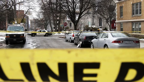 Yellow crime scene tape in sharp focus with urban street scene and parked vehicles blurred in background.