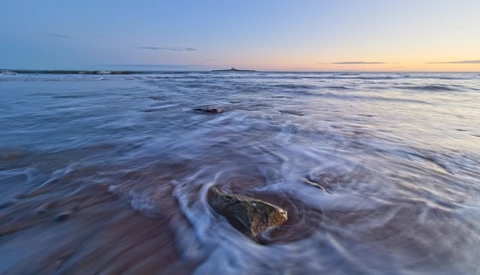 Rocky outcrop surrounded by flowing seawater during twilight, with soft waves creating circular patterns around the stone.