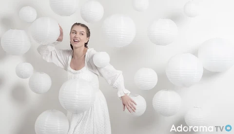 Pregnant woman in white dress posing among floating white paper lanterns against a white backdrop.