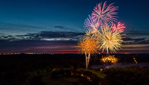 Colorful fireworks burst against a twilight sky above a darkened landscape with scattered lights.