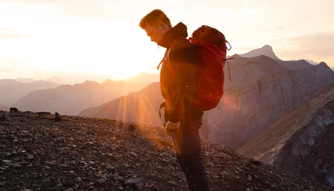 Backpacker with red pack standing on mountainous terrain at sunset, silhouetted against golden light and distant peaks.