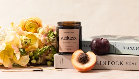 Styled flat lay with yellow flowers, cosmetic jar, sliced peach, dark plum, and stacked books on neutral background.