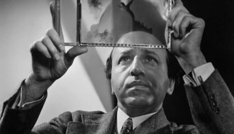 Man in suit holding up a mirror reflecting his face in black and white studio portrait.