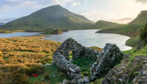 Rugged mountain landscape with cone-shaped peak, glacial lakes, and ancient stone ruins in foreground.