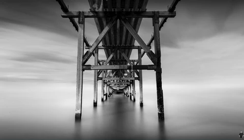 Symmetrical black and white photograph taken underneath a pier, showing repeating wooden support structures receding into a misty distance.