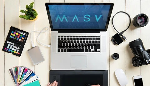 Overhead view of a photographer's workspace with laptop displaying MASV software, camera equipment, and color reference tools.