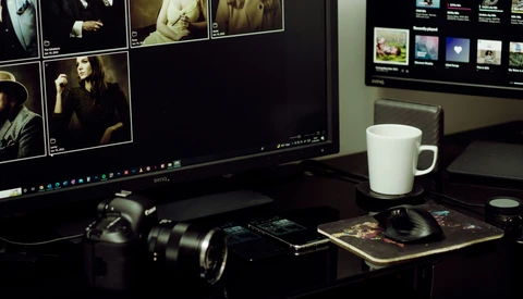 Photographer's desk setup with DSLR camera, lens, and monitor displaying photo editing software.