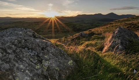 Sunrise over rolling hills with large boulders in foreground and distant mountains.