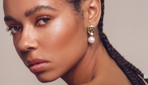 Close-up portrait of a woman with defined eyebrows and warm-toned makeup, wearing pearl drop earrings.