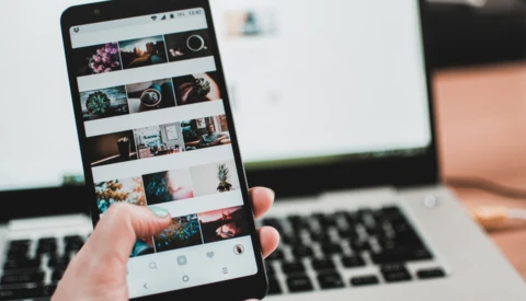 Smartphone displaying a grid of colorful photographs in a gallery app, held in hand above a laptop.