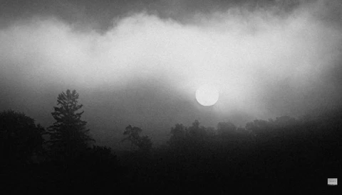 Sun obscured by heavy storm clouds above a dark forested landscape at dusk or dawn.