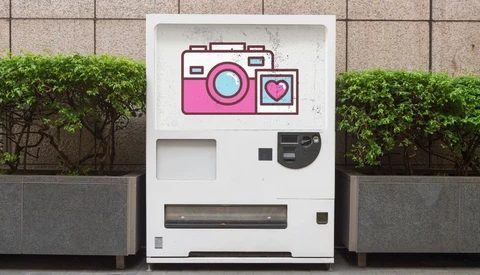 Illustration of a pink and white instant camera vending machine with a heart icon on its display.