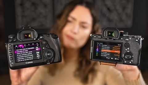 Two Canon mirrorless cameras held up side by side, displaying rear LCD screens with menu interfaces and settings visible.