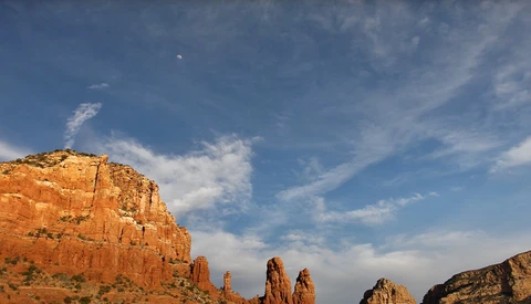 Desert rock formations with towering red sandstone cliffs against a blue sky with wispy clouds.