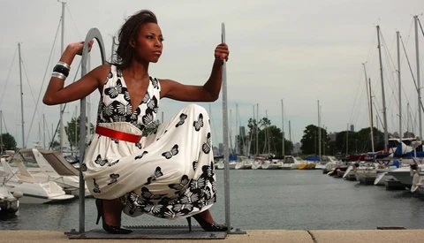 Woman in patterned dress posing with rope at a marina lined with sailboats.