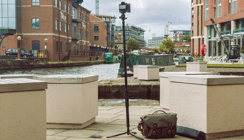 Camera on tripod positioned at waterfront location with brick buildings and modern development in background.