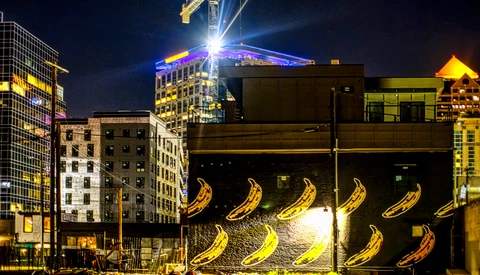 Nighttime urban street with illuminated banana murals on dark building facade surrounded by lit skyscrapers.