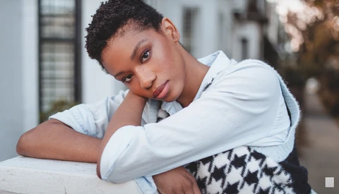 Young woman with short hair resting her head on her hand, wearing a light blue shirt and patterned vest.