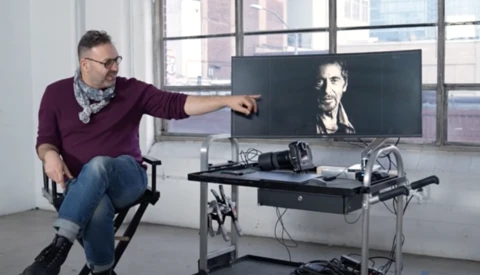 Man in burgundy sweater points at portrait displayed on wide monitor at editing workstation.