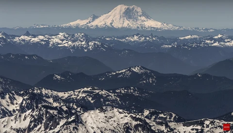 Snow-capped volcanic peak rising above layered mountain ranges in distant haze.