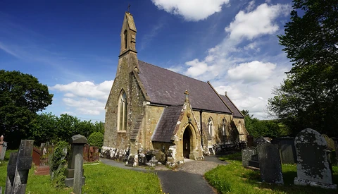 Stone chapel with steep pitched roof and tall spire surrounded by weathered gravestones in overgrown cemetery.