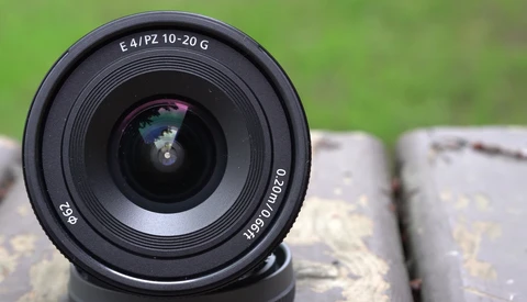Sony E 10-20mm f/4 PZ G wide-angle zoom lens viewed head-on, mounted on brick surface with blurred green background.