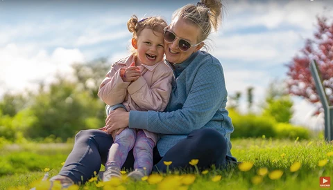 Woman and young child sitting together on grass, both wearing sunglasses and smiling at camera.