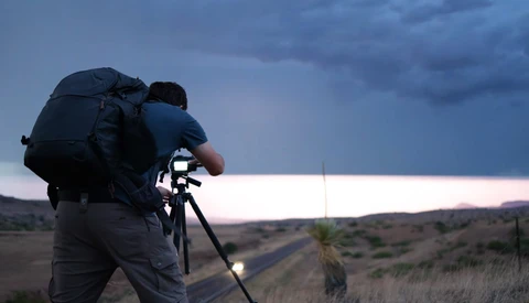 Photographer with large backpack operating a telephoto lens on tripod at dusk overlooking an expansive landscape.