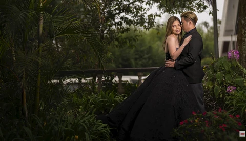 Man in dark suit embracing woman in black ball gown in lush garden setting with flowering plants.