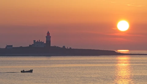 Silhouetted lighthouse and coastal buildings on a peninsula at golden hour sunset.