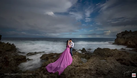 Couple embracing on rocky beach in flowing pink gown with long exposure ocean and dramatic sky.