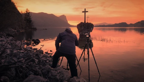 Photographer operating a camera on tripod at sunrise beside a calm lake with mountains and a distant cross silhouetted against orange sky.