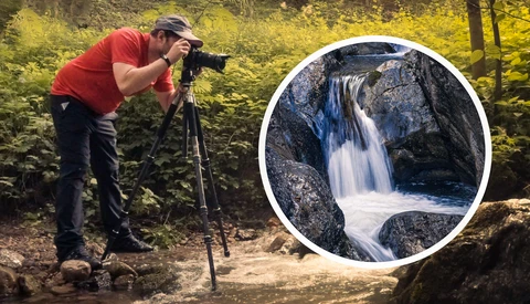 Photographer in red shirt and cap photographing a cascading waterfall in a forested area using a tripod-mounted camera.