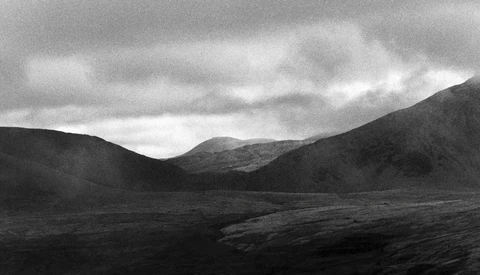 Mountainous valley landscape under dramatic cloudy sky in black and white.