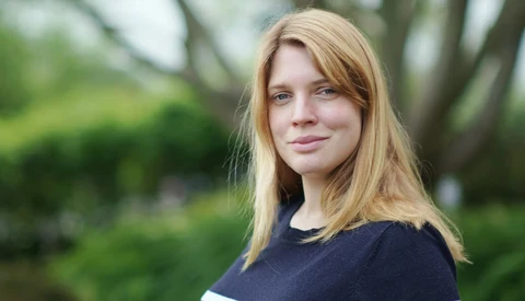 Woman with strawberry blonde hair looking toward camera in outdoor setting with blurred green foliage background.