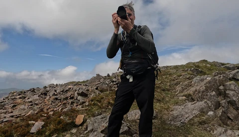 Photographer holding camera up to face while standing on rocky hillside with sparse vegetation.