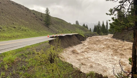 Swollen river with muddy water rushing past a damaged highway embankment during flood conditions.
