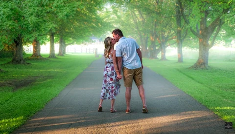 Couple kissing on a tree-lined path with bright sunlight filtering through green foliage.