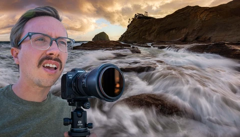 Photographer with camera on tripod at rocky coastline with dramatic waves and sunset sky.