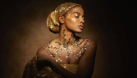 Woman with gold leaf makeup and glittering body art against warm brown background.