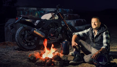 Man crouching by a campfire next to a motorcycle at night.