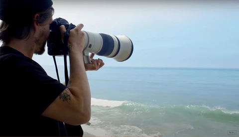 Photographer holding telephoto lens while shooting coastal ocean and beach landscape.